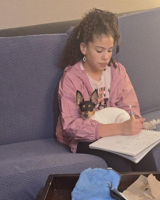 Ava Silva sitting on a couch with a small dog in her lap. She is holding a pen and writing on a piece of paper. 
