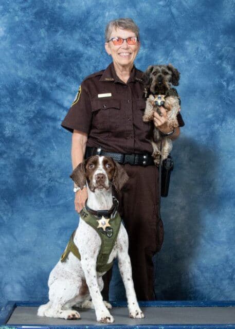 Sue Stejskal, wearing a police uniform is holding two dogs.