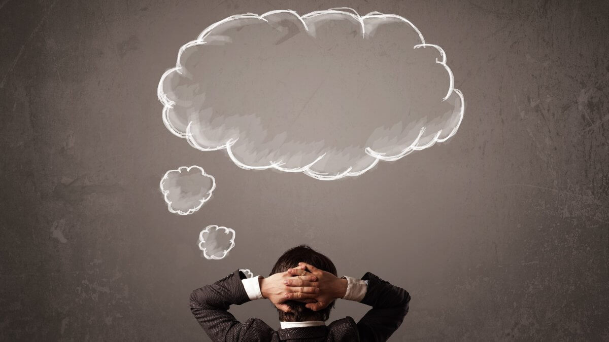 Businessman sitting with cloud thought above his head. Young businessman sitting in office chair in front of a wall with cloud thought sketched on a chalkboard above his head