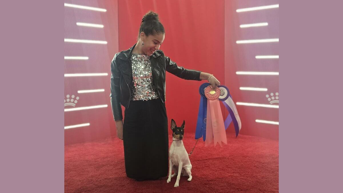 Ava Silva kneeling on a red carpet holding a blue ribbon. A small dog is standing next to her.