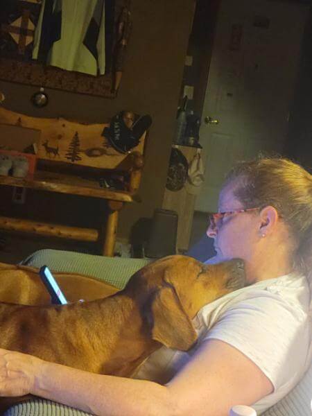 Julie Buss is seated, staring at her phone, with her sleeping puppy, Mirabel, curled up on her lap.