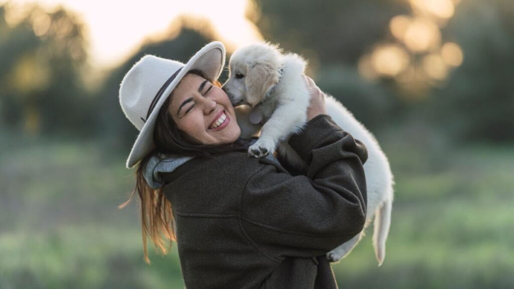 Young Caucasian woman wearing a hat standing in grassy field, lovingly holding Golden Retriever puppy.