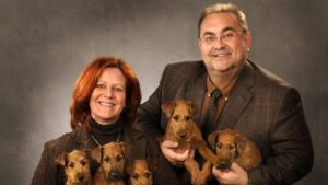 Photo of a mana and woman on grey background, holding Irish Terrier puppies.