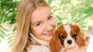 Portrait happy blonde girl with long hair holding cute Purebred puppy Cavalier King Charles Spaniel. Tree leaves on summer morning in light outdoors sun with natural blurry background, close-up.