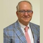 David Fitzpatrick, an older man, one of the judges, wearing glasses, a blue plaid suit jacket, a white shirt, and a light pink tie stands in front of a plain light-colored wall.