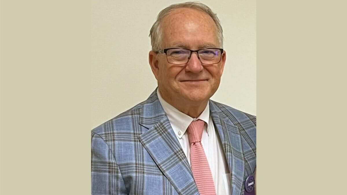 David Fitzpatrick, an older man, one of the judges, wearing glasses, a blue plaid suit jacket, a white shirt, and a light pink tie stands in front of a plain light-colored wall.