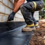 Construction worker waterproofing building foundation with bituminous membrane, protecting against moisture and leaks. Construction worker applying bituminous waterproofing membrane to building foundation, sealing against potential water damage and structural deterioration AI generated