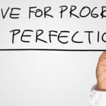 Man writing the idiom - Strive For Progress Not Perfection - on a virtual interface with a black marker pen over grey with copyspace in a motivational business message.