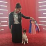 Ava Silva kneeling on a red carpet holding a blue ribbon. A small dog is standing next to her.